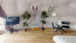 A tented accommodation with a TV, two potted plants, a swing, a table with chairs, and a hanging light fixture.