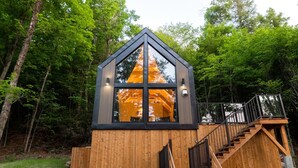 A wooden cabin with a triangular roof, a staircase, and a window reflecting a tree. - Bel Air Tremblant CALAFATE : 1 CHAMBRE, JACUZZI, SAUNA (La Conception)