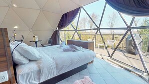 Ein modernes Schlafzimmer mit einem großen Bett, einem Nachttisch, einer Lampe und Blick durch große Fenster.