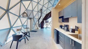 Une cuisine moderne dotée d’un plafond en verre aux motifs géométriques, un coin repas avec une table et des chaises en verre, et un comptoir de cuisine équipé d’appareils électroménagers et d’ustensiles.