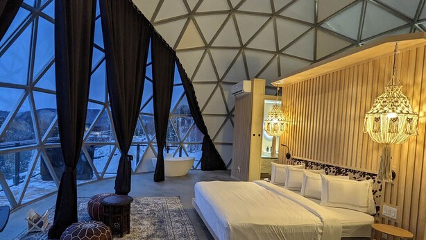Une chambre dotée d’un grand lit, d’une baignoire et offrant une vue sur un paysage enneigé grâce à une fenêtre à structure géométrique.