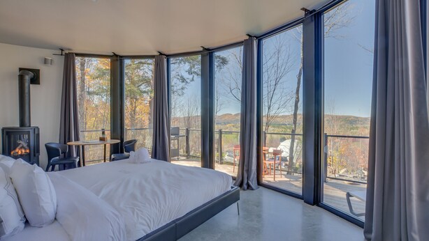 A bedroom with a large bed, a fireplace, and a balcony with a view of trees and hills. - Bel Air Tremblant ROUND LOFT : 1 LIT KING, JACUZZI ET SAUNA (La Conception)