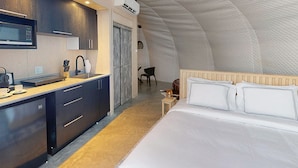 A compact hotel room with a kitchenette, a bed with white linens, and a curved ceiling.