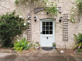 Un edificio de piedra con una puerta blanca, celosía de madera y un pequeño jardín con flores.
