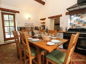 Un comedor con una mesa de madera dispuesta para cuatro personas, sillas con cojines verdes, una cocina con salpicadero de azulejos y un ventanal con vistas a la vegetación.