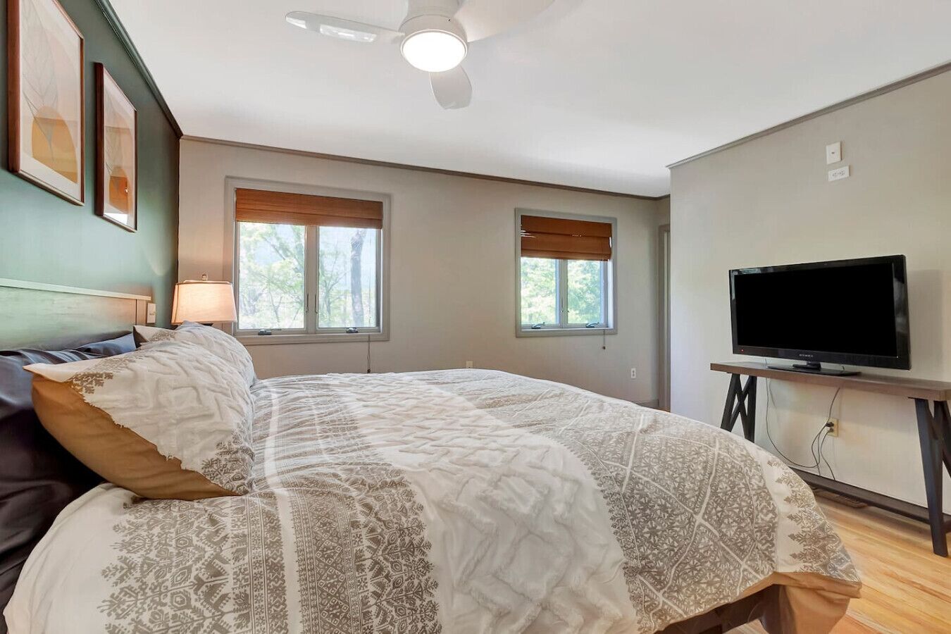 A bedroom with a bed, two windows with blinds, a ceiling fan, and a TV on a stand.