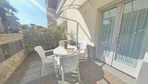 A small outdoor patio with a round table and white plastic chairs, a potted plant, and a sliding glass door. - Cozy Nest in a Great Area (Lignano Sabbiadoro)