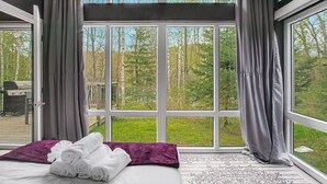 Un dormitorio con un ventanal grande, una cama con ropa blanca, un plaid morado y una toalla enrollada.