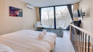 Una habitación de hotel moderna con una cama grande, un área de descanso con una silla y una mesita, un televisor montado en la pared y vistas a árboles por la ventana.