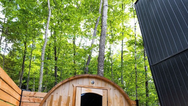 Estructura de sauna de madera con puerta, rodeada por una terraza de madera y un área boscosa.