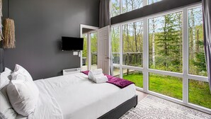 Ein modernes Schlafzimmer mit einem großen Bett, einem Flachbildfernseher und Blick auf eine Terrasse und Bäume.