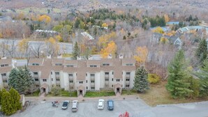 A multi-story building with a parking lot, surrounded by autumn-colored trees and a hilly landscape in the background. - Stratton walk on/off, walk to food/drink, fireplace (Stratton)
