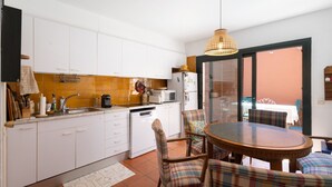 Una cocina con armarios blancos, un comedor con mesa redonda y sillas a cuadros, y vistas a través de un ventanal.