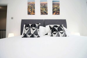Un lit agrémenté d’oreillers aux motifs géométriques, une tête de lit grise et trois photographies de paysages urbains accrochées au mur.