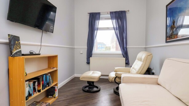 A room with a sofa, a chair, a bookshelf, a flat-screen TV, a window with curtains, and a radiator. - Bartletts Base (Stockton-on-Tees)