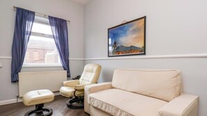 Une pièce avec un canapé beige, un fauteuil blanc, un fauteuil de bureau noir, un radiateur, une fenêtre avec des rideaux bleus et un tableau encadré accroché au mur.