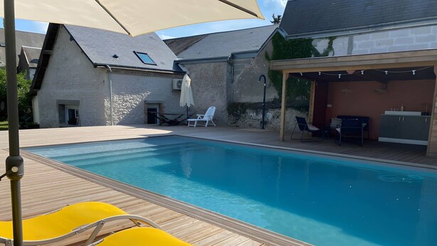 Ein Swimmingpool mit Holzterrasse und gelben Liegestühlen, ein überdachter Patio-Bereich und ein Gebäude aus Stein im Hintergrund.