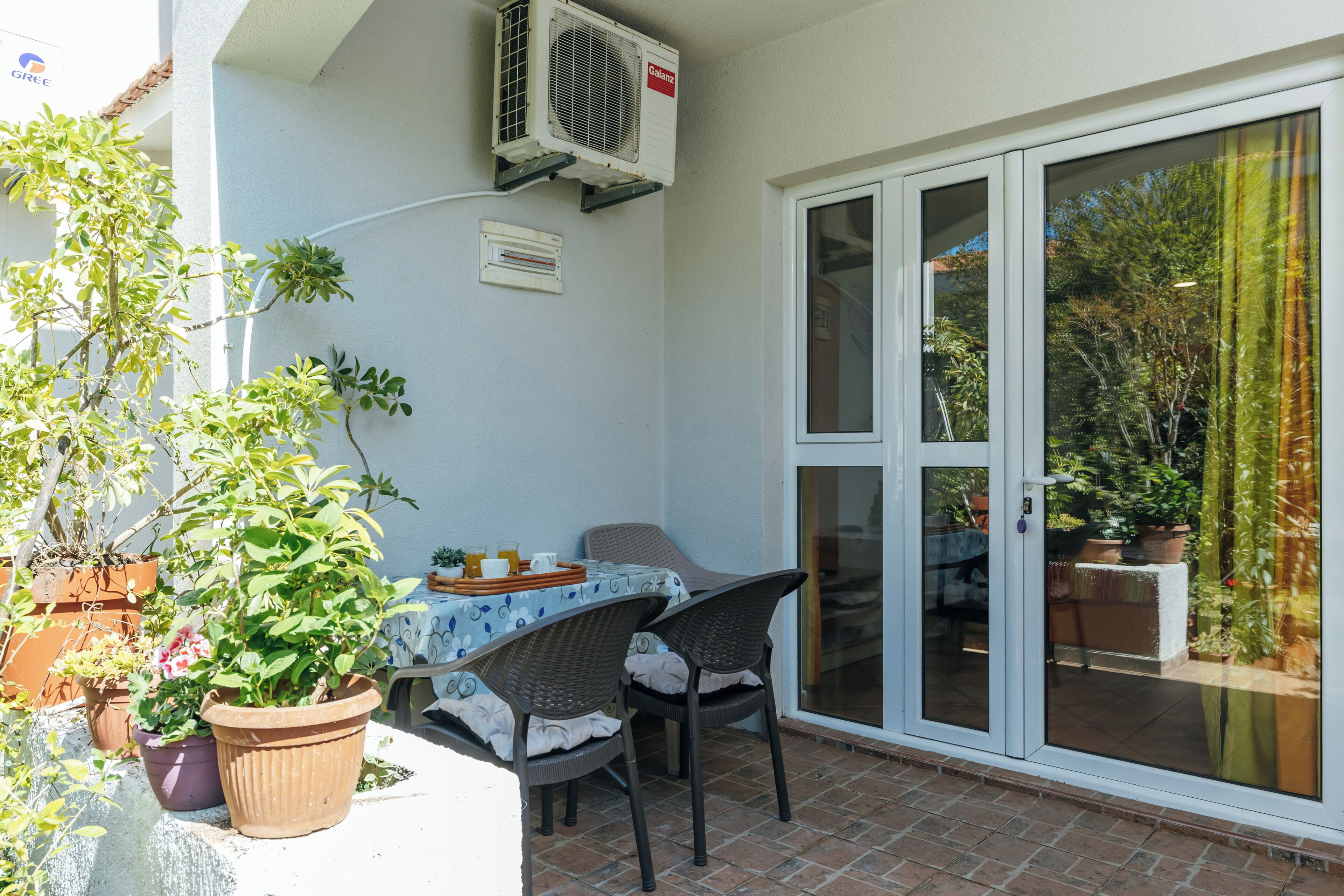 Une terrasse avec une table et des chaises, des plantes en pot, un appareil de climatisation et une porte-fenêtre coulissante donnant sur un jardin.