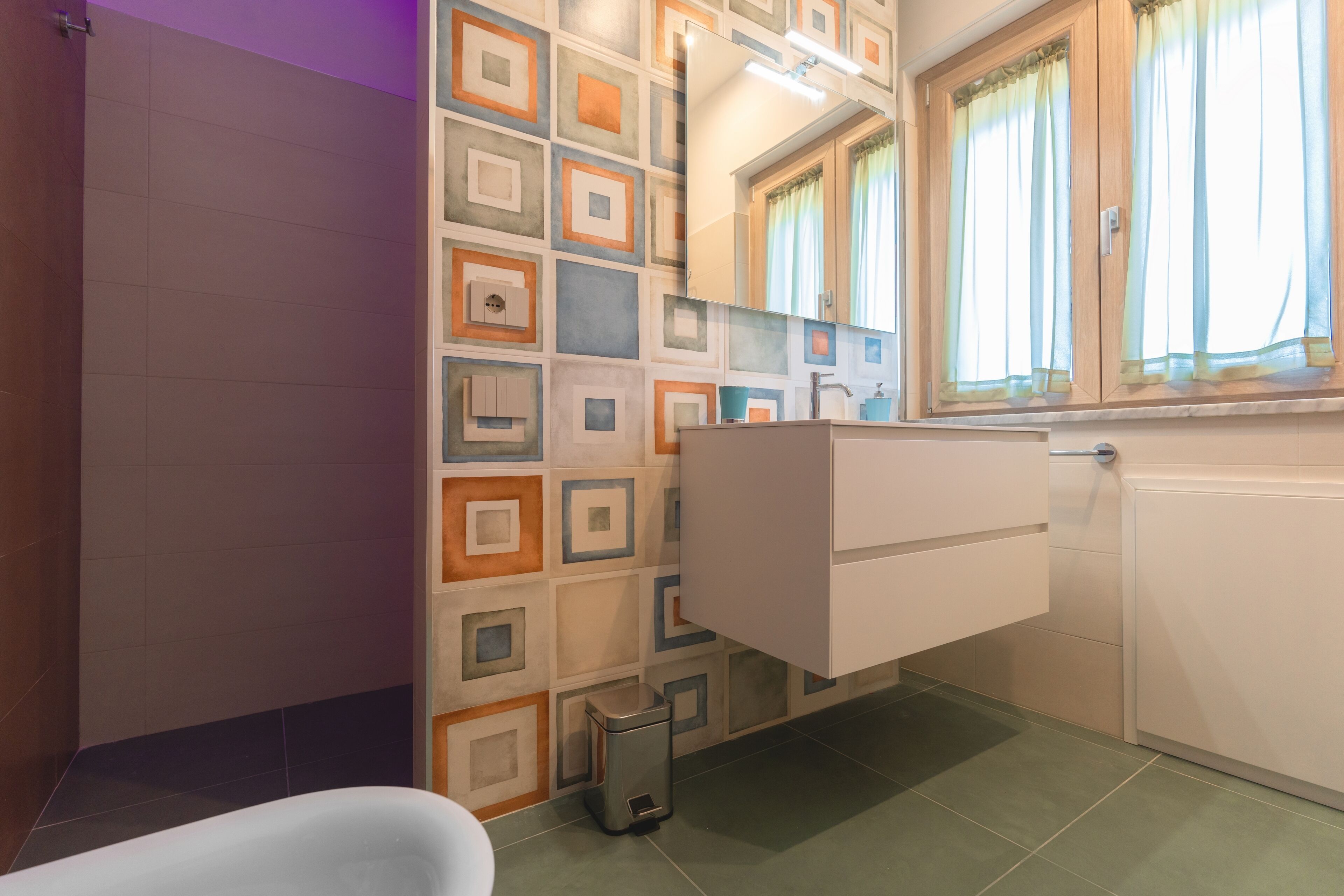 A bathroom with a patterned wall, a sink, a mirror, and a window with curtains.