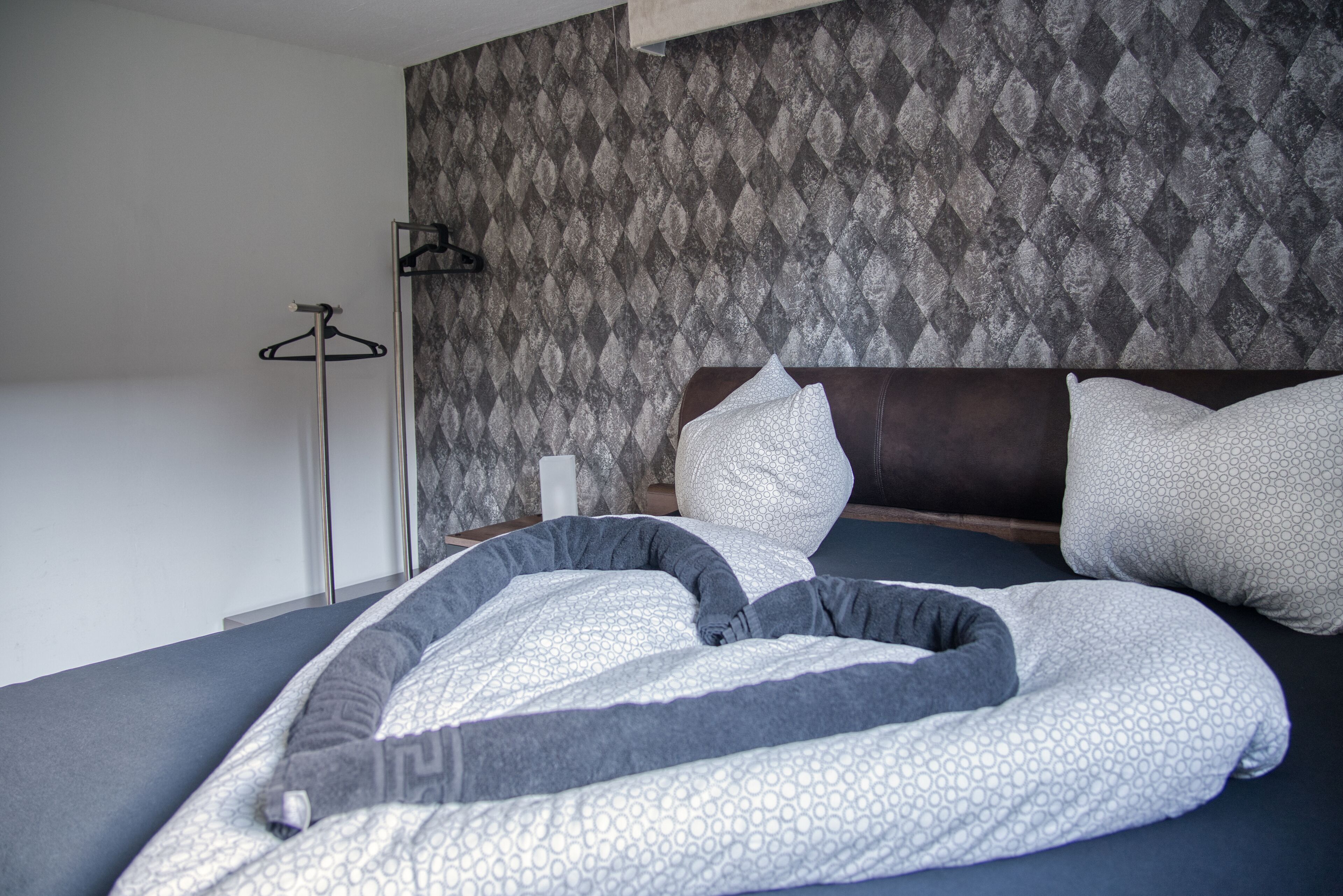 Een net opgemaakt bed met een beddengoed met een patroon, een hoofdbord en een wand met behang in ruitmotief.