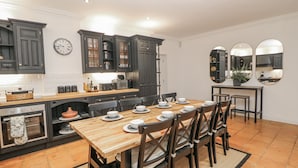 A modern kitchen with dark cabinetry, a wooden dining table set for six, and a large mirror above the counter. - Hallfield Hall Cottage (Alfreton)