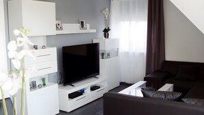 A modern living room with a flat-screen TV, white entertainment unit, dark sofa, and a large window with curtains.