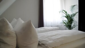 A hotel room with a bed, pillows, a plant, and sheer curtains.