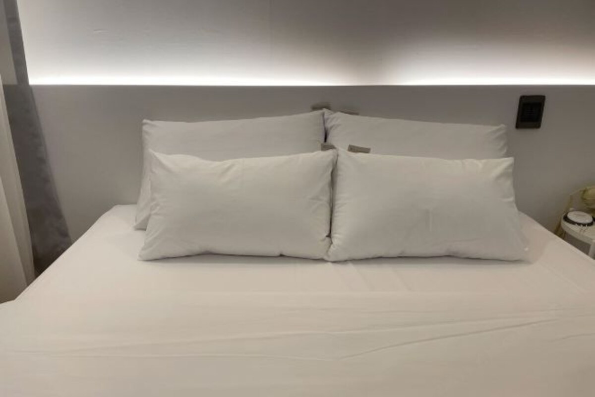 A neatly made bed with white linens and pillows, a bedside table with a lamp, and a wall-mounted light fixture.