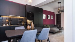 A modern kitchen with dark cabinetry, a dining table with chairs, and a living area with a sofa and a red accent wall. - Premium Designer Retreat in Central Athens Gizi (Athina)