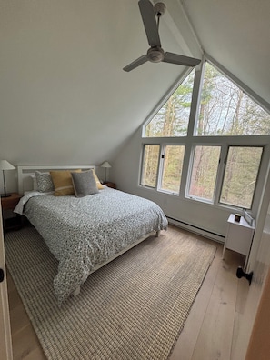 Quarto com cama, ventilador de teto, janela grande e vista para árvores.