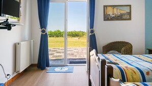 Une chambre à coucher comprenant un lit, un téléviseur, un fauteuil en osier et une vue sur un espace verdoyant, visible à travers une porte coulissante.