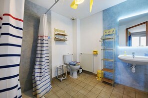 A bathroom with a toilet, a sink, a shower curtain, a shelf with toiletries, and a radiator. - “Vol de mouettes” Guest Room with Shared Garden and Wi-Fi (Huisnes-sur-Mer)