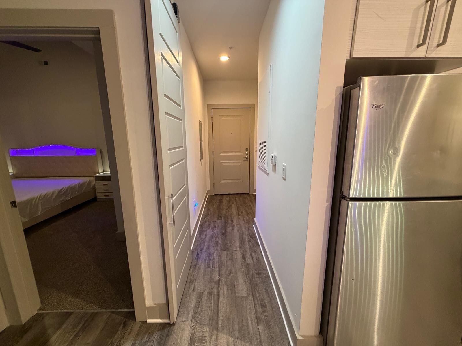 Un pasillo estrecho con una cama, un refrigerador y una puerta que da a otra habitación.