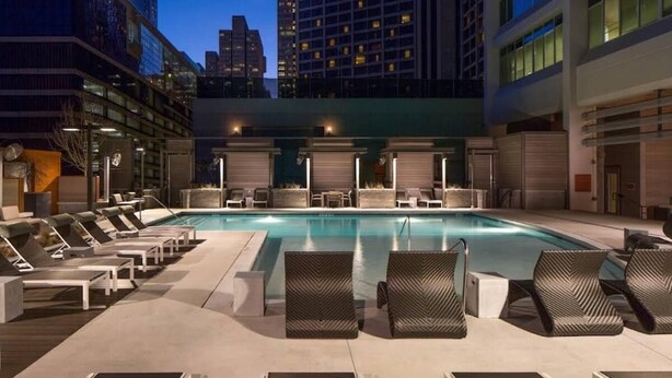 Una piscina en la azotea con sillones de descanso, vista a edificios de la ciudad y cielo despejado.