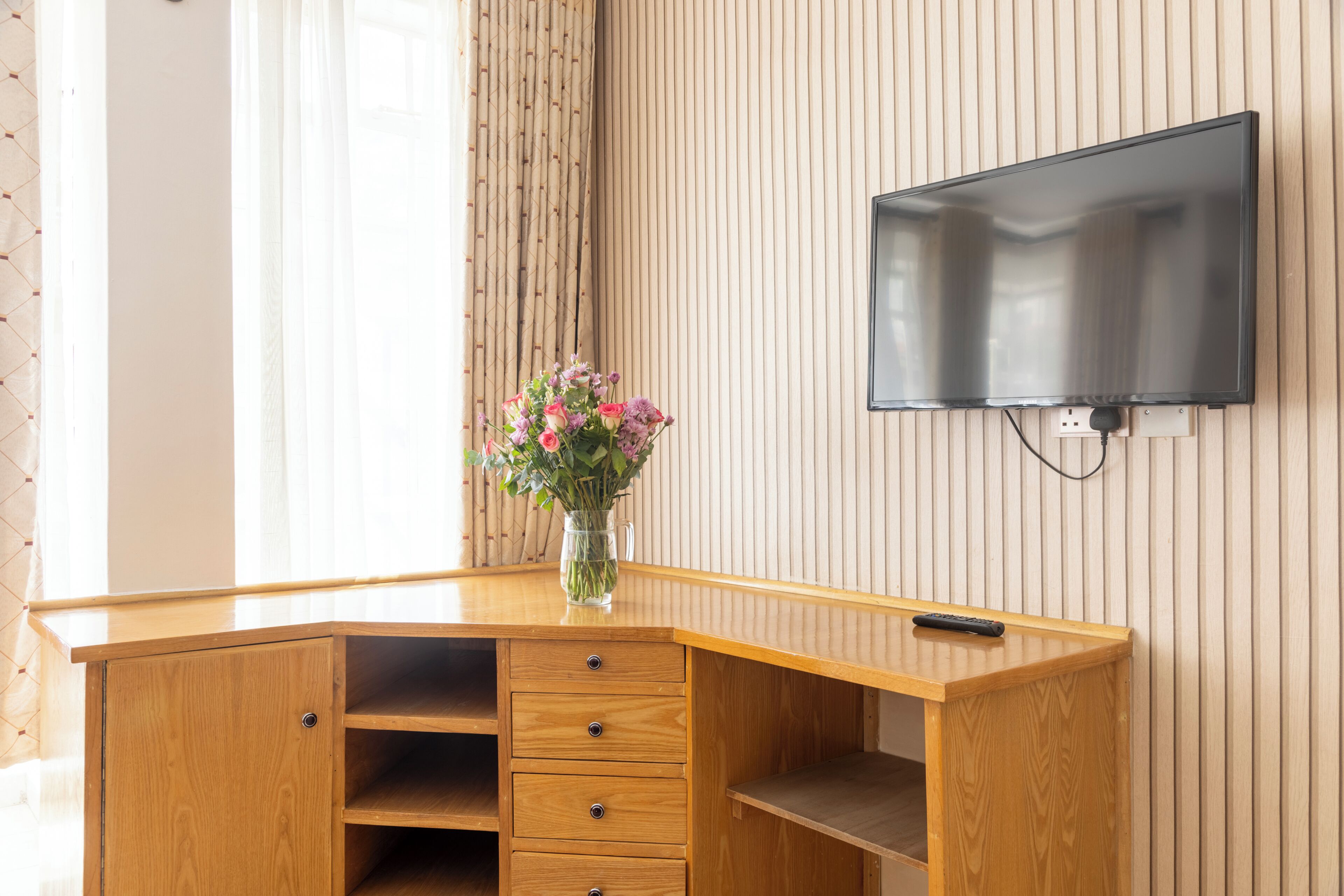 Un bureau en bois orné d’un vase de fleurs, un téléviseur fixé au mur et une télécommande posée sur le bureau.