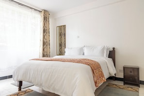 A hotel room with a bed, a nightstand, a mirror, and a window with curtains. - Gemina Court  (Nairobi)