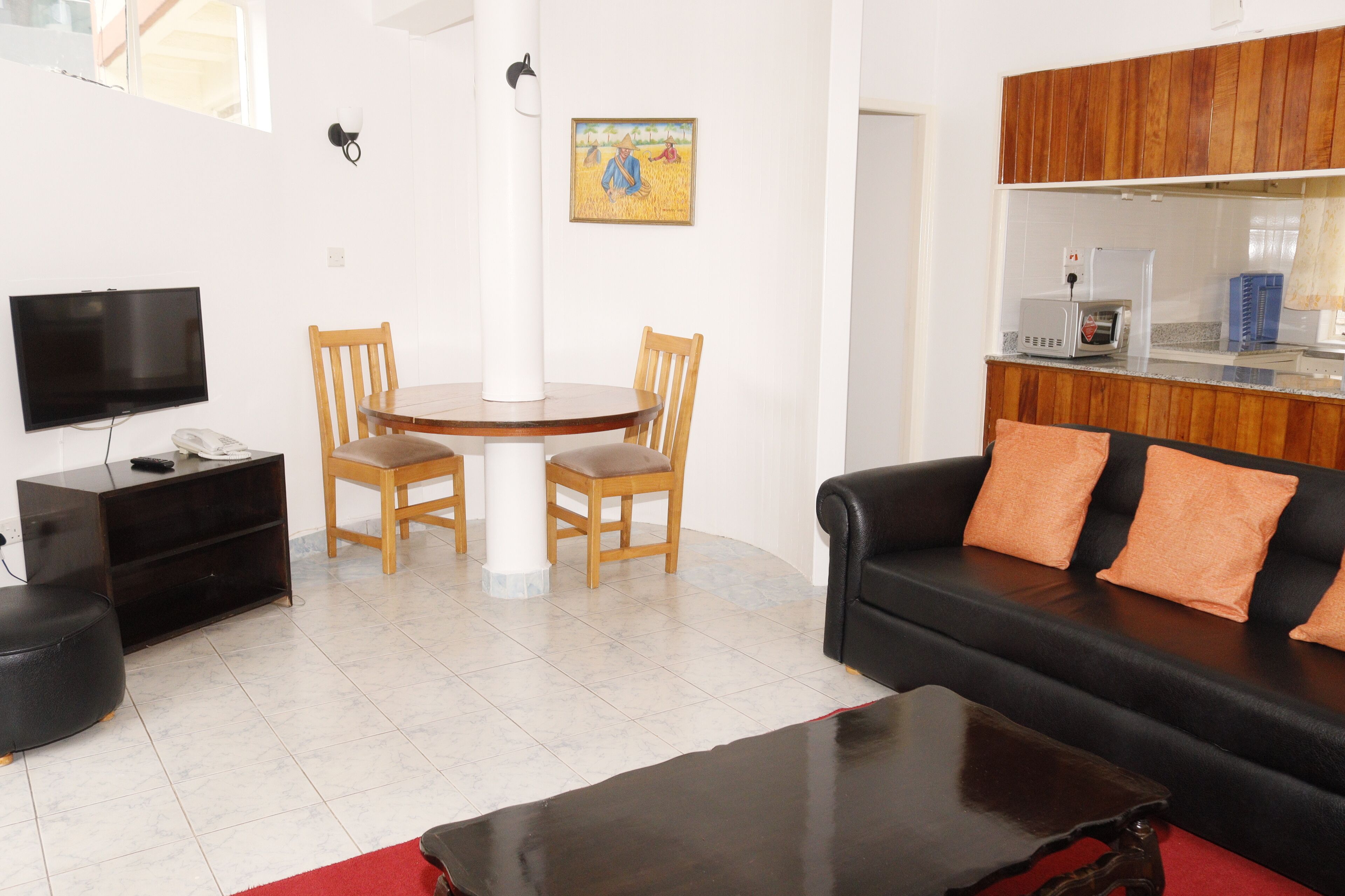 A living room with a black sofa, wooden chairs, a TV, a dining table, and a kitchen area with wooden cabinets.