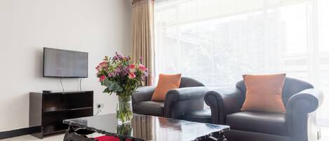 A modern living room with a flat-screen TV, a glass vase with flowers, two leather sofas with orange cushions, and a red carpet.