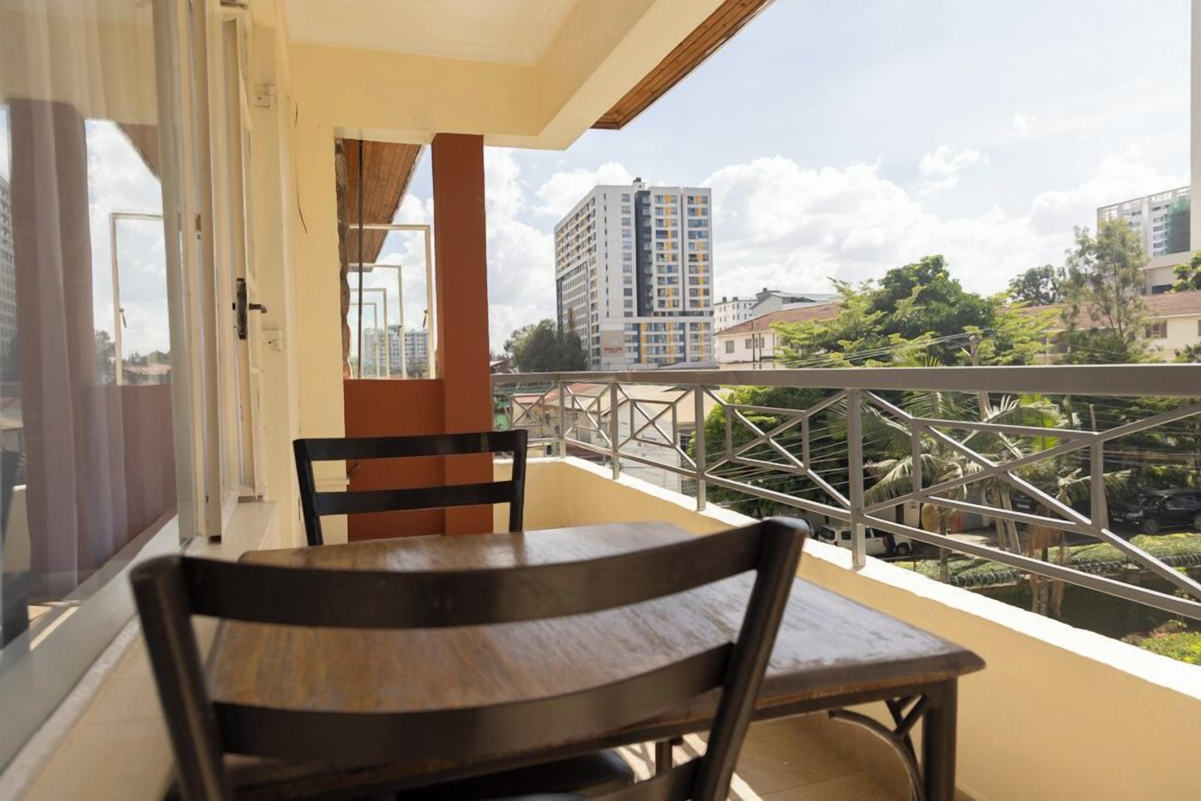 A balcony with a wooden table and chairs, overlooking a cityscape with buildings and greenery.