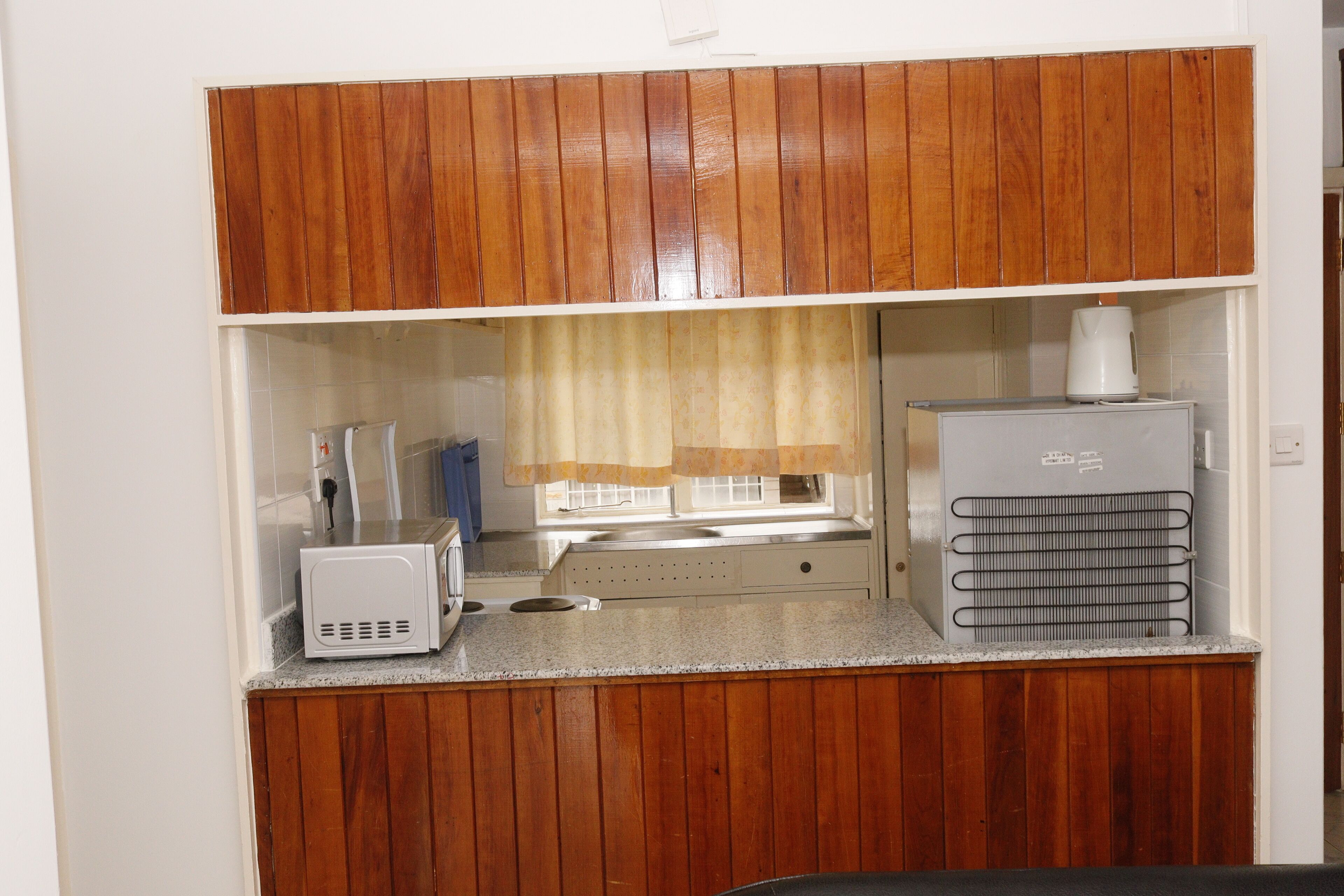 A compact kitchenette with wooden cabinets, a microwave, a kettle, and a water dispenser.
