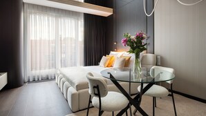 Ein modernes Hotelzimmer mit einem Bett, einem Glastisch, zwei Stühlen, einer Blumenvase und einem großen Fenster mit Vorhängen.