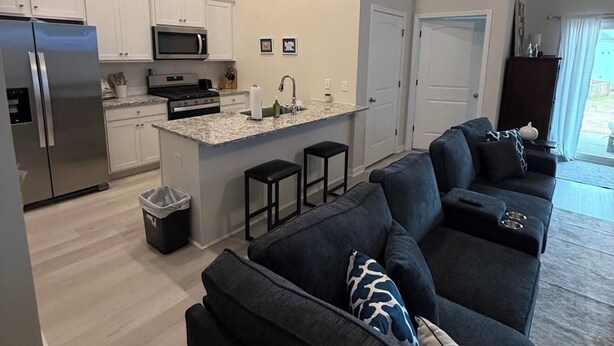 A modern kitchen with white cabinets, a stainless steel refrigerator, and a microwave. There is a dark-colored sofa with a throw pillow and a small coffee table.
