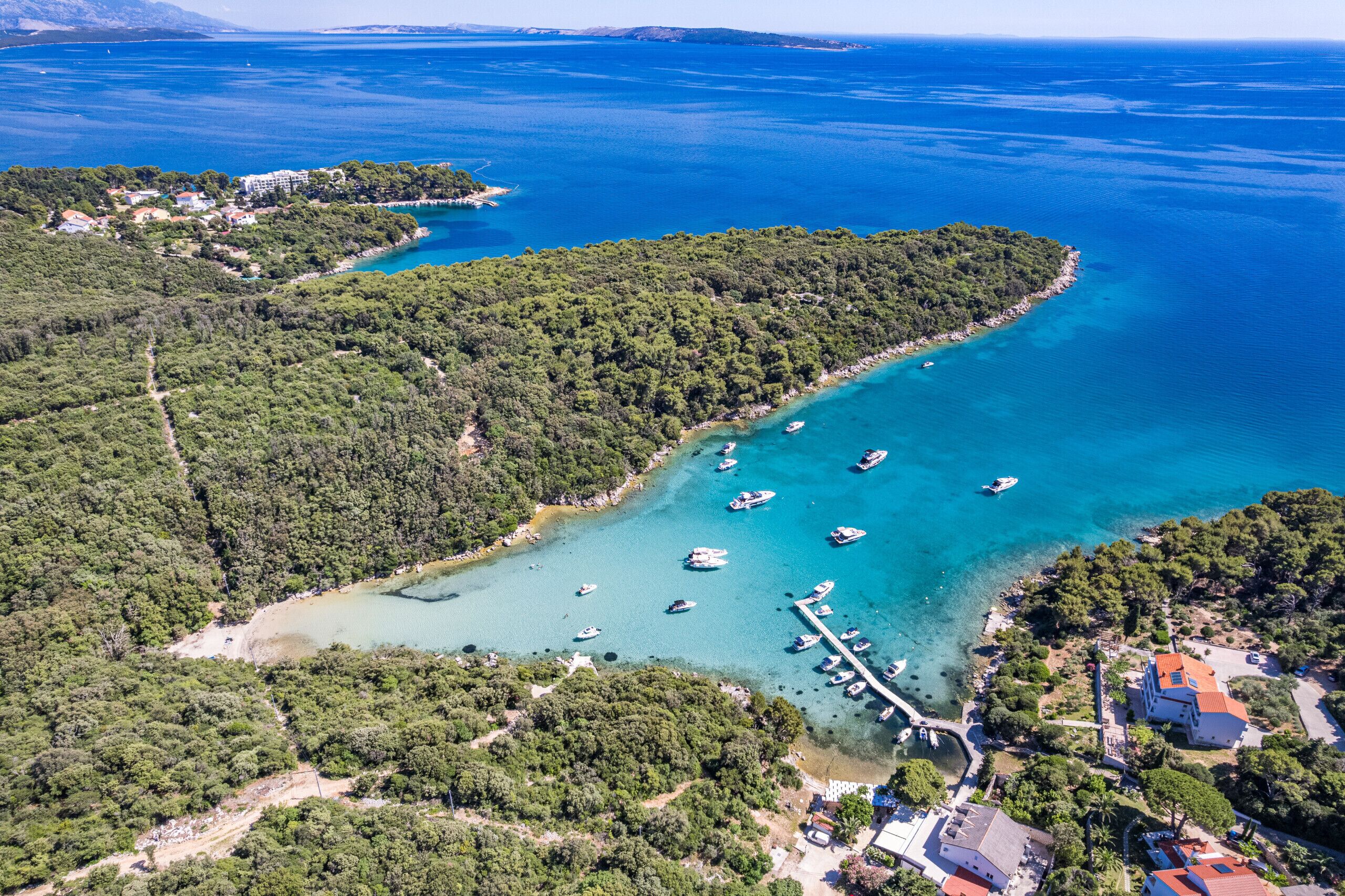 An aerial view of a coastal area with boats docked at a pier, surrounded by dense forest and a clear blue sea.