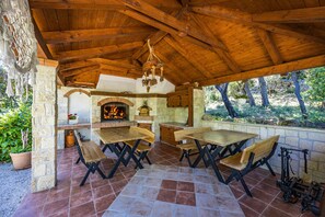 An outdoor patio with wooden tables and benches, a stone fireplace, and a hanging light fixture. - Petra Sea View Apartment 2 (Kampor)