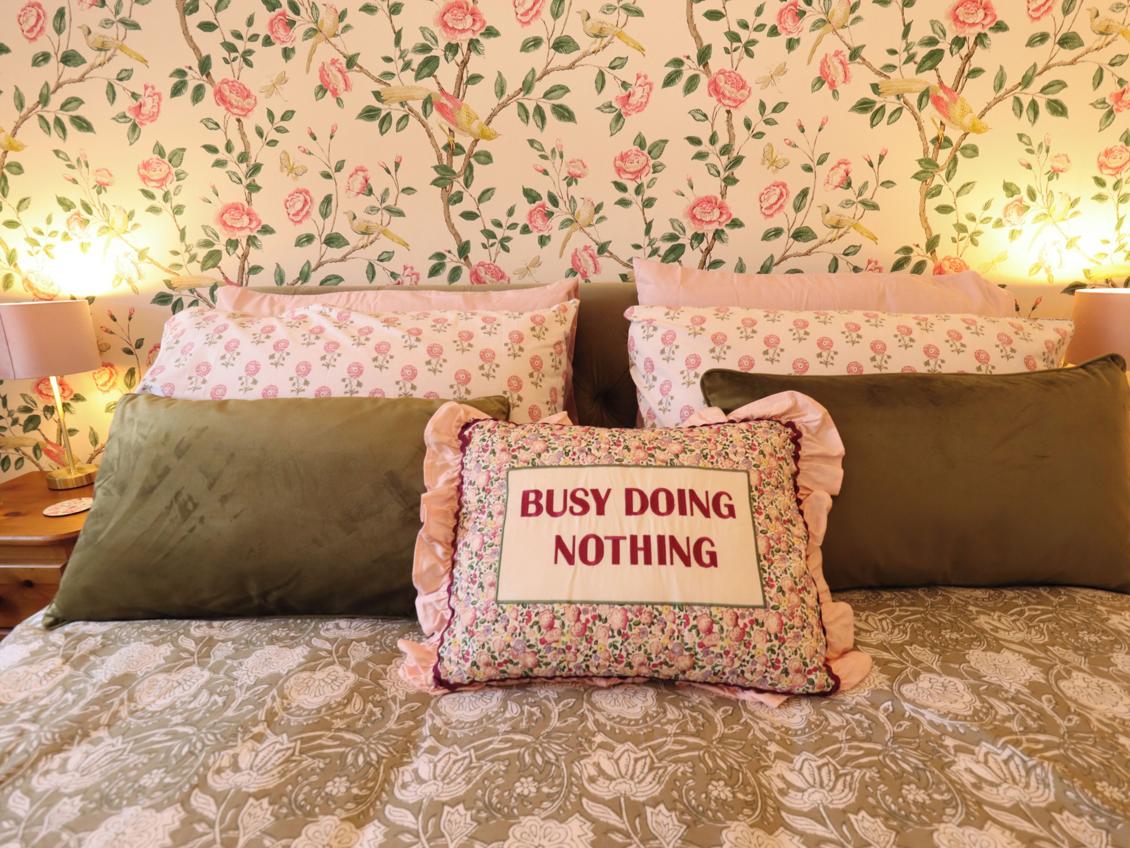 A bed with floral-patterned bedding, a pillow with 'BUSY DOING NOTHING' text, and a wall with a floral wallpaper design.