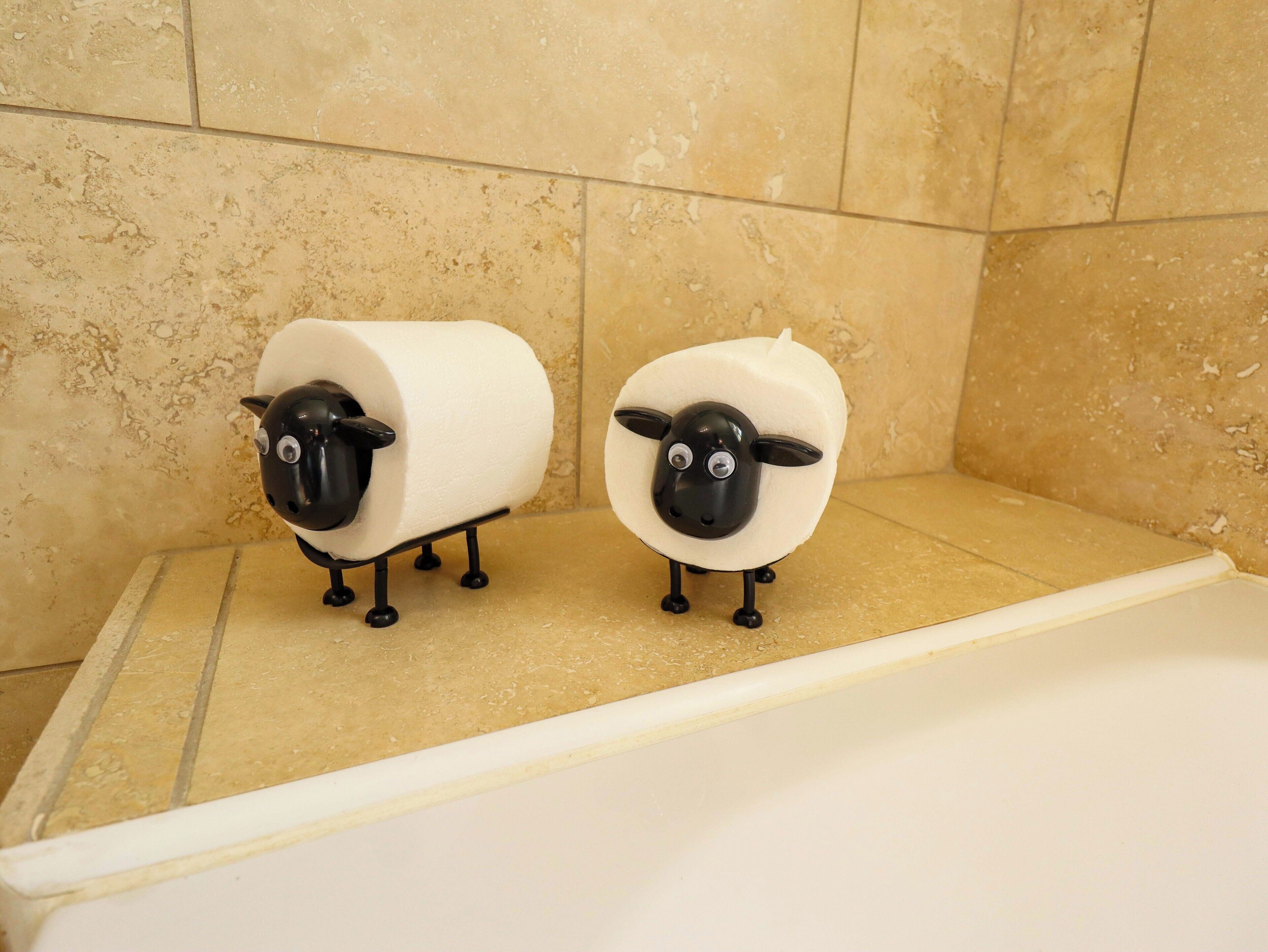 Zwei Toilettenpapierhalter in Form von Schafen in einem Badezimmer mit beige gefliesten Wänden.