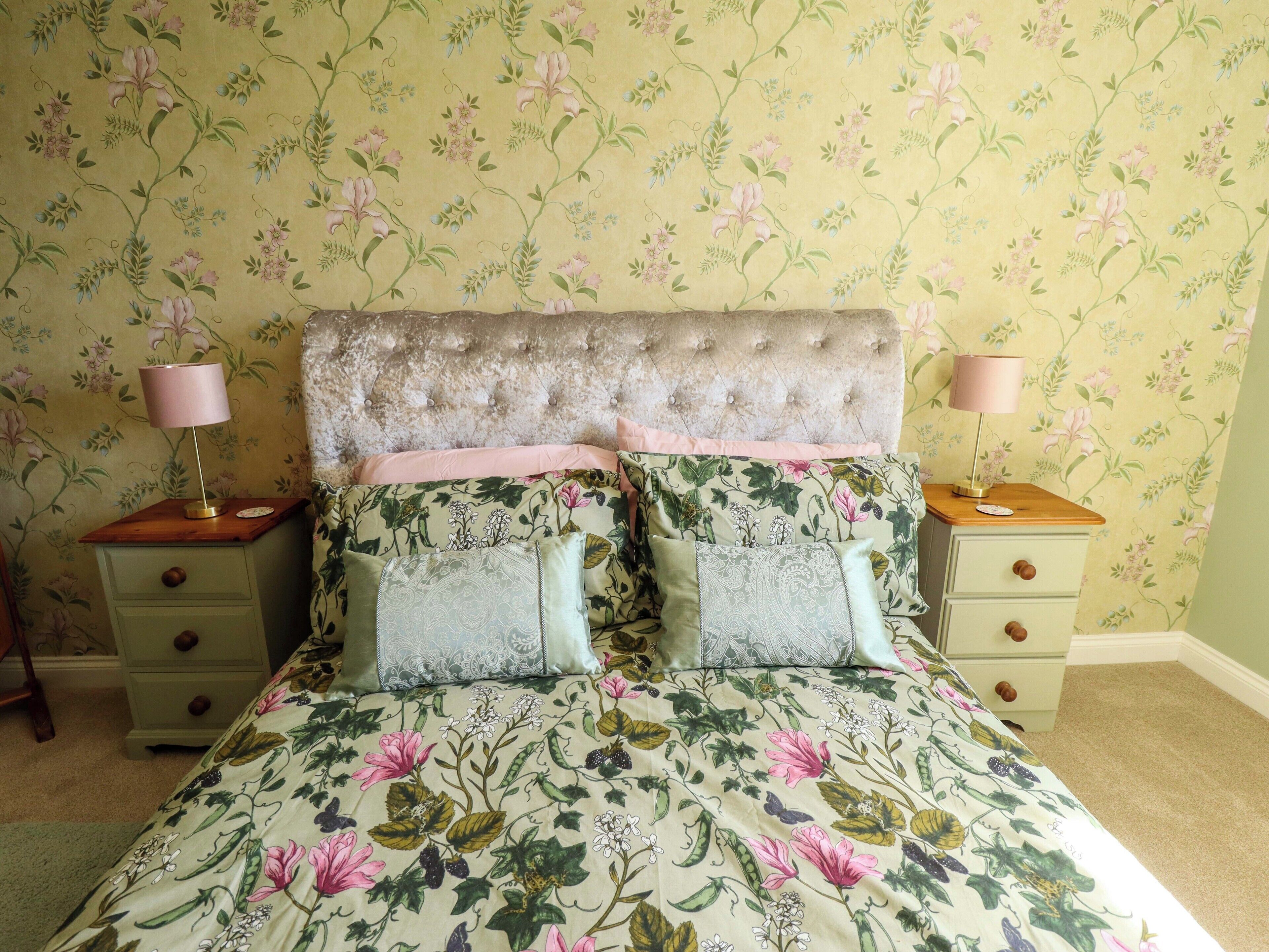 A bedroom with a floral wallpaper, a tufted headboard, two bedside tables with lamps, and a bed with floral bedding.