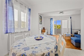 A room with a dining table, chairs, a sofa, a small table with a lamp, and a view through a glass door. - Petra Sea View Apartment 4 (Kampor)