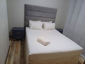 Ein ordentlich bezogenes Bett mit grauem Kopfteil, weißen Kissen und einem aufgerollten weißen Handtuch.