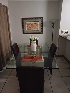 Ein Essbereich mit einem Glastisch, zwei Stühlen, einer weißen Vase mit Blumen, einem gerahmten Bild an der Wand und einer Stehlampe.